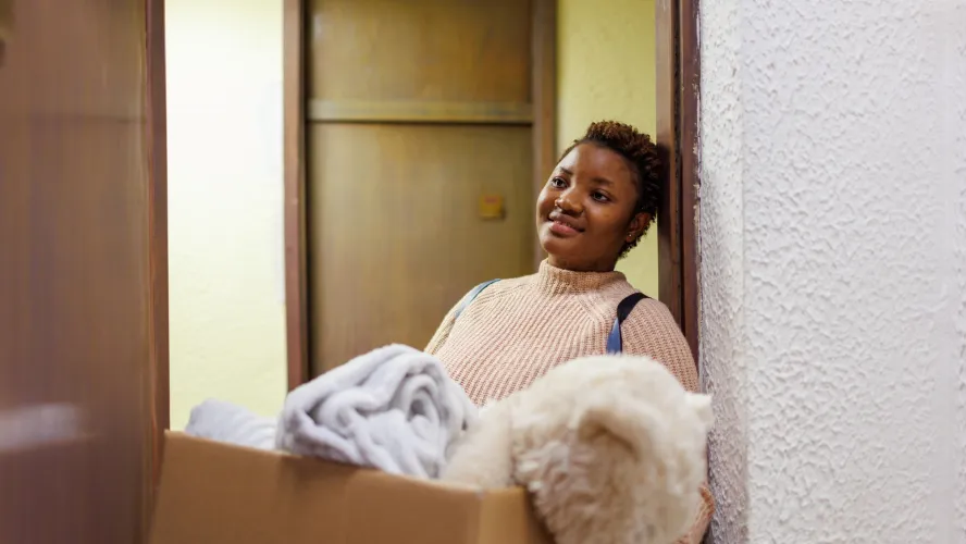 Woman carrying a box of household items, smiling indoors.
