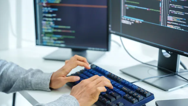 Hands typing on keyboard with dual monitors displaying code