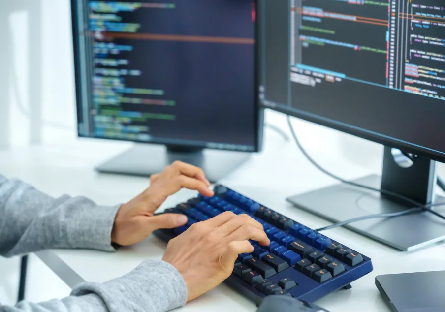 Hands typing on keyboard with dual monitors displaying code