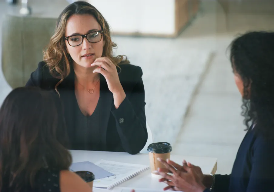 Three women discussing at a table with documents and coffee cups
