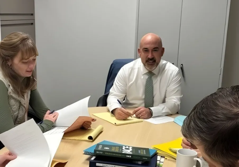 CM Fellow Bryan Farrell smiles while seated at a table with court staff—a woman and a man—during a meeting. Papers are spread across the table as they collaborate.