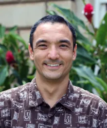 Man wearing brown patterned shirt smiling outdoors with greenery background.
